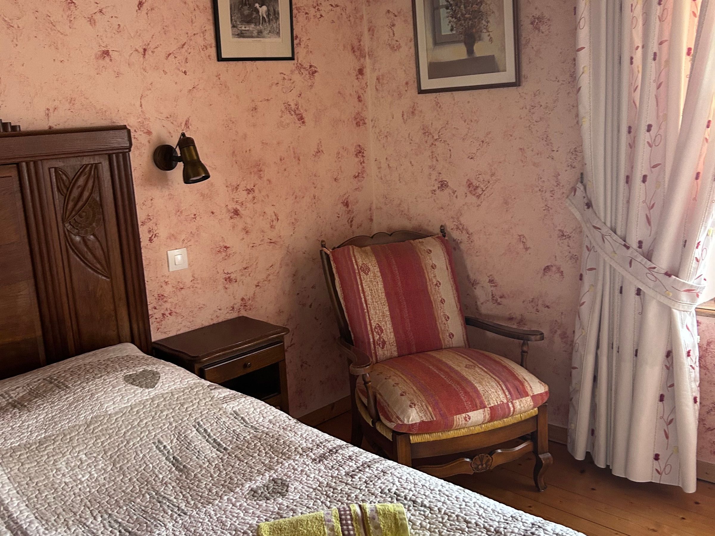 Une chambre à coucher comprenant un lit, une chaise, une table de chevet, une lampe fixée au mur et un rideau.