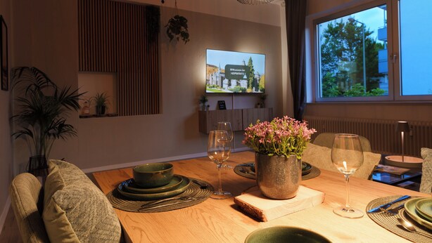 A dining area with a wooden table set for two, a television mounted on the wall, and a window with a view of trees. - Castora Homes - City Apartment Kassel near train station (Kassel)