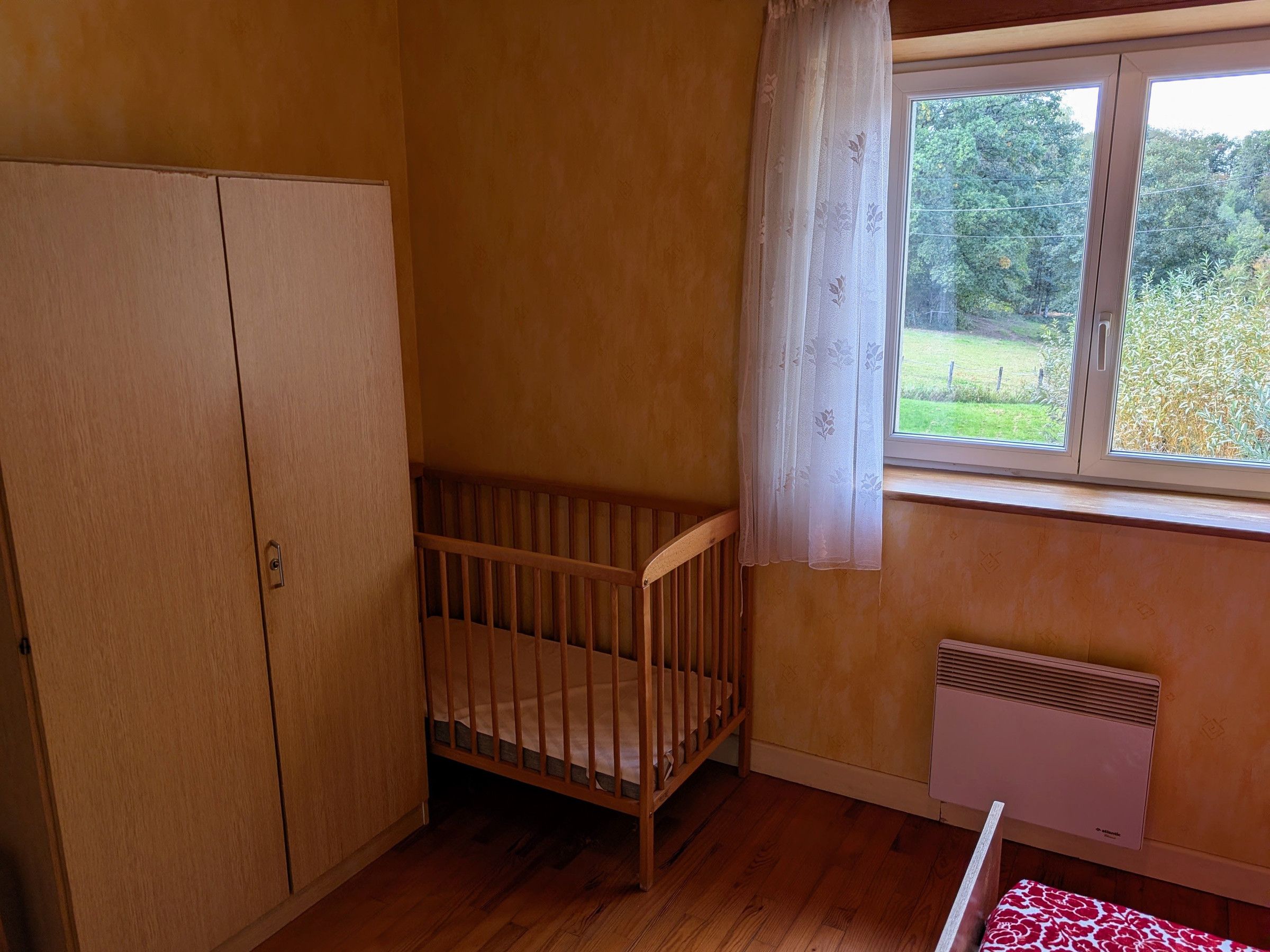 A room with a wooden crib, a wardrobe, a radiator, and a window with a view of greenery.