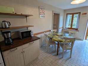 A small kitchen with a microwave, kettle, and coffee maker on the counter. There is a table with chairs and a window with curtains.