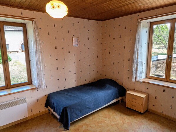 A small, single-bed room with a wooden ceiling, a window on one side, and a small bedside table.