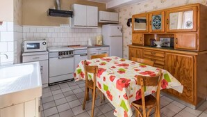 Een kleine keuken met witte kasten, een magnetron, een fornuis en een spoelbak. Er staat een houten eettafel met stoelen.