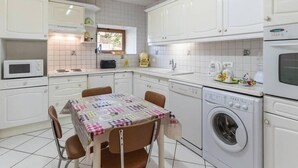 Uma cozinha compacta com armários brancos, micro-ondas, máquina de café, máquina de lavar e uma mesa com toalha xadrez.