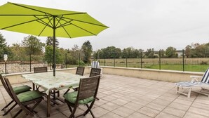 Um pátio externo com mesa e cadeiras, um guarda-sol verde e vista para um campo cercado.