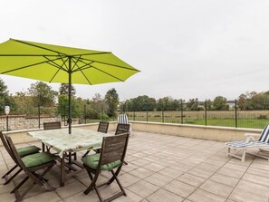 Eine Terrasse im Freien mit Tisch und Stühlen, einem grünen Sonnenschirm und Blick auf ein eingezäuntes Feld.