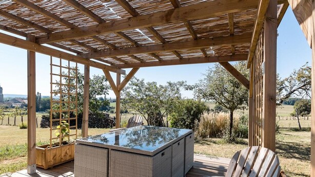 Une terrasse en bois avec un jacuzzi à débordement en verre, des meubles en osier et une chaise en bois.