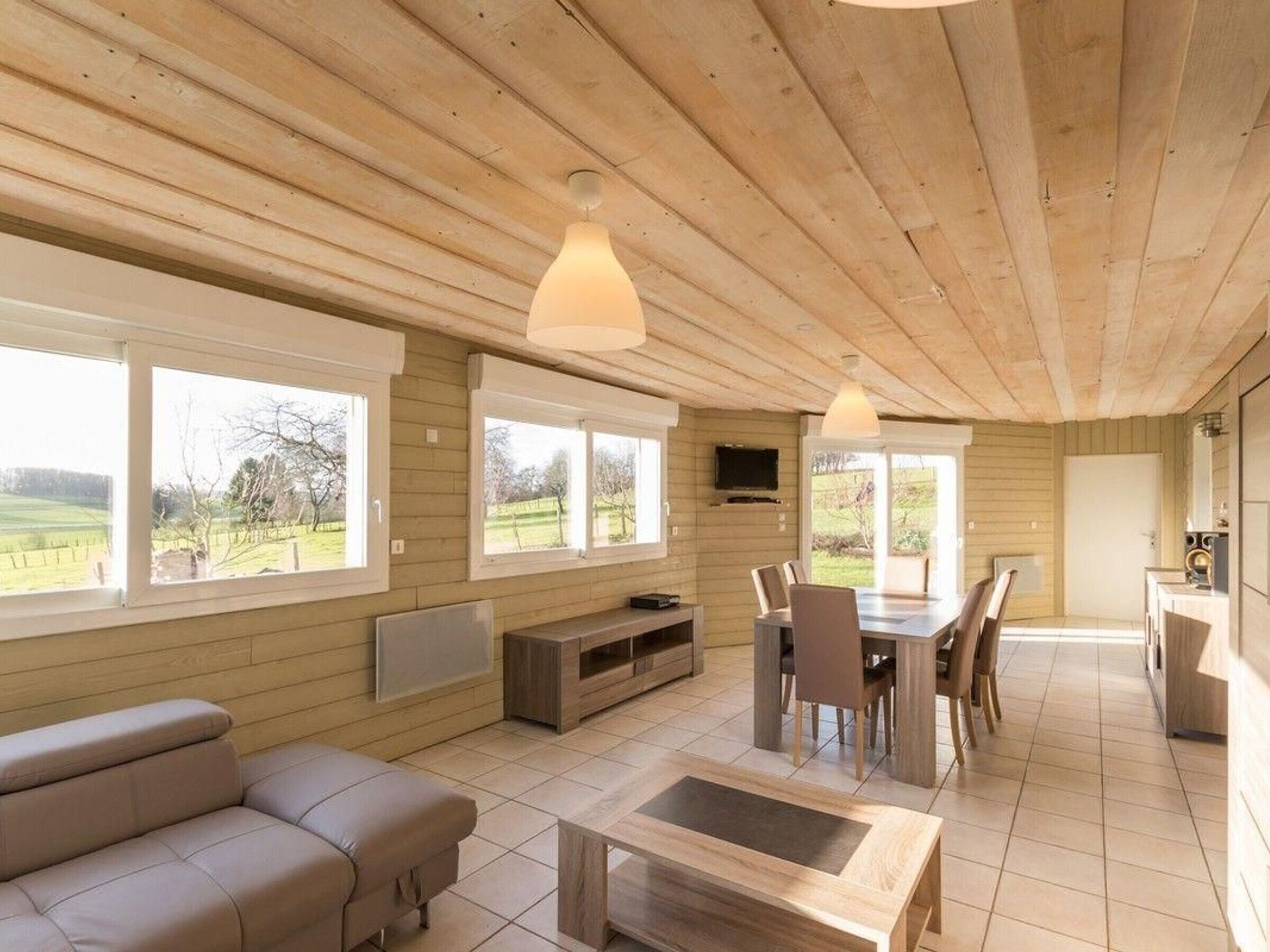 A spacious living area with a sofa, dining table, and chairs, large windows offering a view of the outdoors, and a television mounted on the wall.
