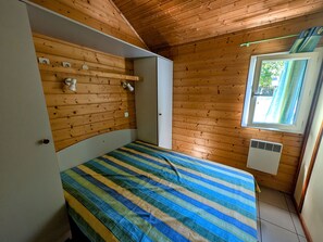 Una habitación pequeña con cama, paredes de madera, una ventana y un conducto de aire blanco.