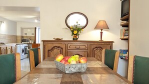 Un comedor con mesa de madera, sillas tapizadas en verde, una cesta de frutas, un aparador de madera con espejo y una lámpara.