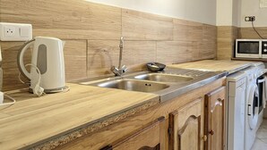 Una cocina con armarios de madera, un lavamanos de acero inoxidable, una tetera, un microondas y una lavadora.