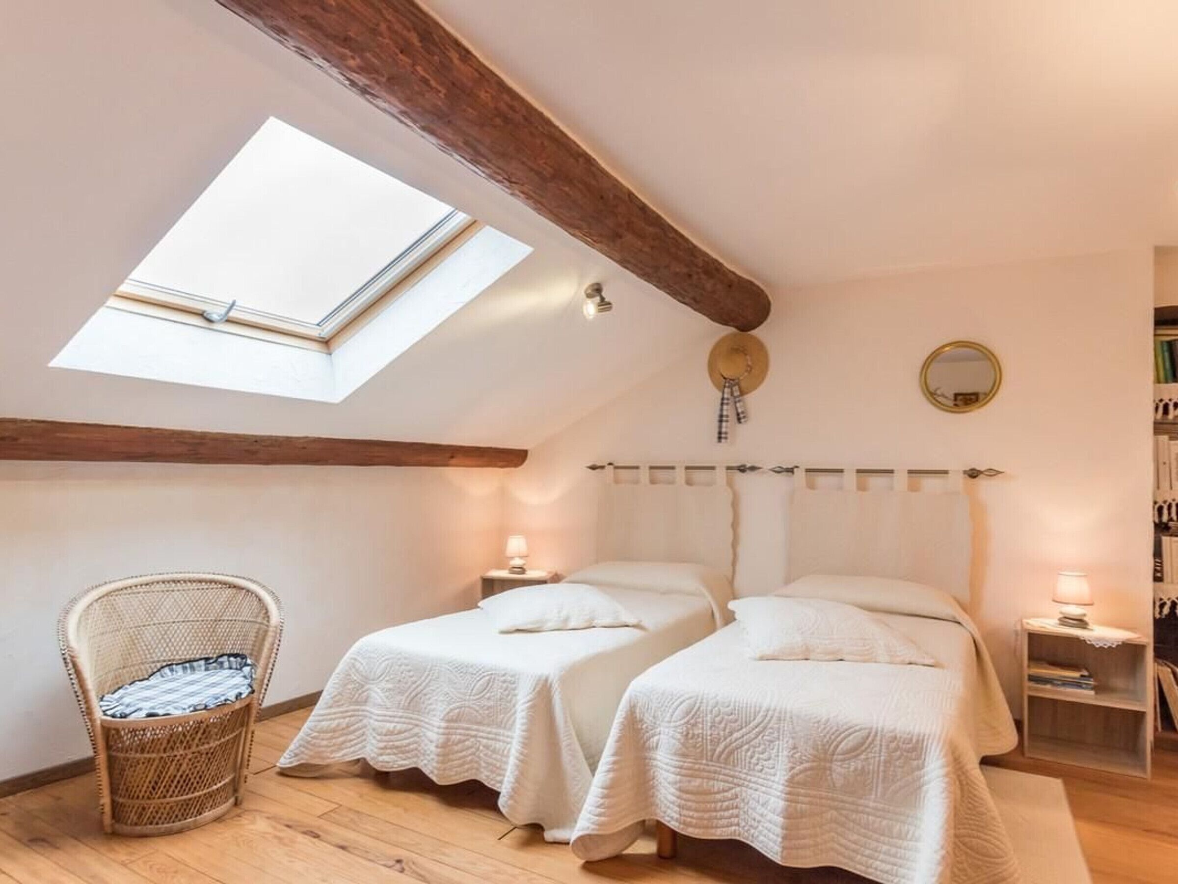 A cozy bedroom with two beds, a skylight, a wicker chair, bedside tables with lamps, and a bookshelf.