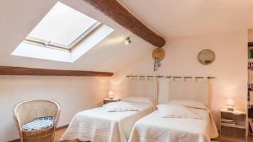 A cozy bedroom with two beds, a skylight, a wicker chair, bedside tables with lamps, and a bookshelf.