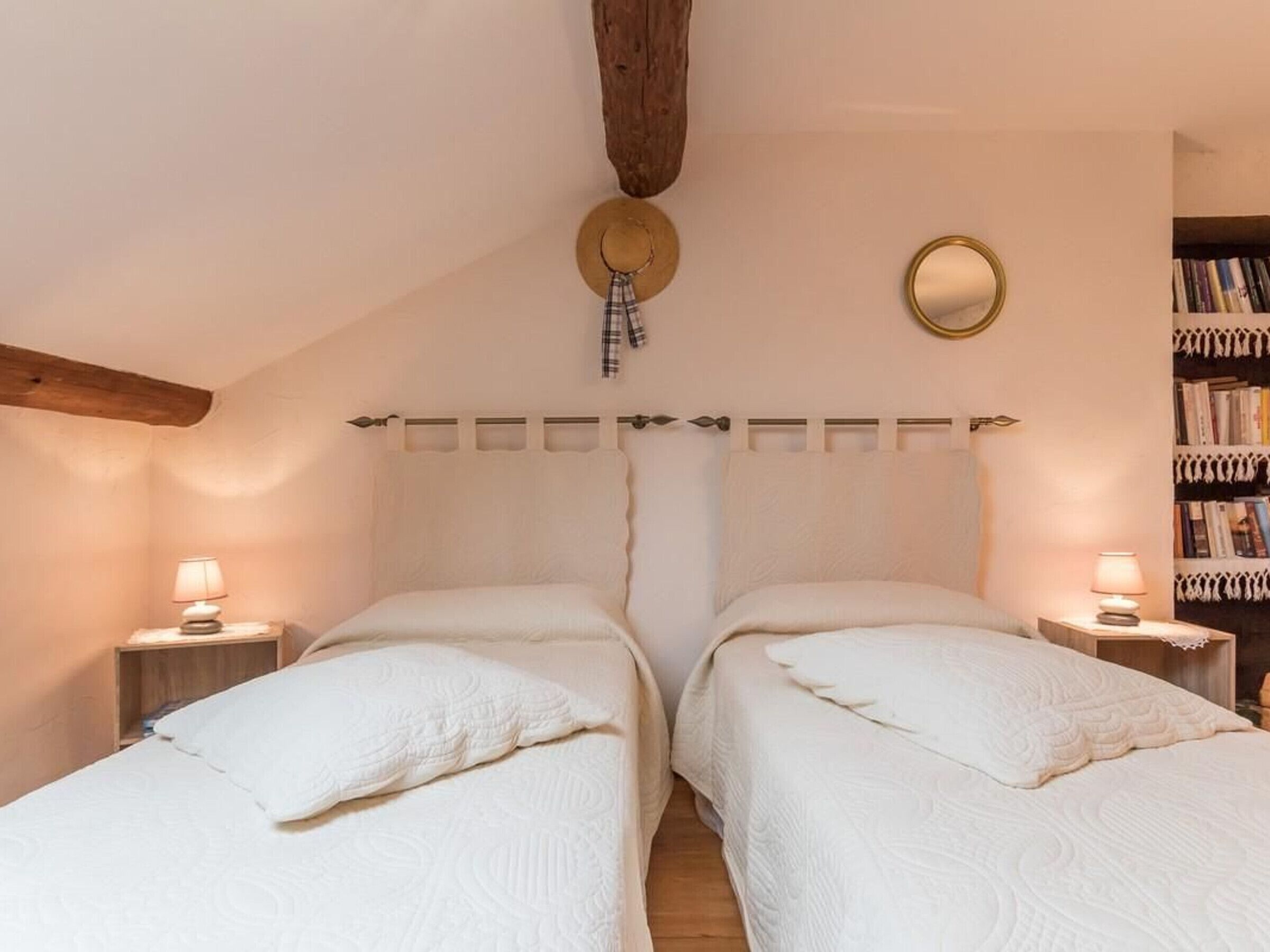 Une chambre avec deux lits, une poutre en bois au plafond, un miroir rond et une bibliothèque.