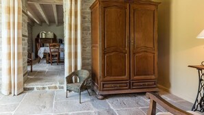 A stone-floored room with a wooden wardrobe, a wicker chair, and a table with a lamp.