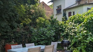 Patio esterno con tavolo e sedie, un barbecue e rigogliosa vegetazione.
