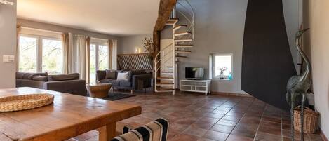 A spacious living room with a wooden dining table, striped chairs, a sofa set, a TV on a white stand, and a spiral staircase.