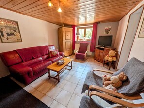 Un salon chaleureux avec un canapé en cuir rouge, une table basse en bois, un fauteuil gris, un téléviseur et un ours en peluche.