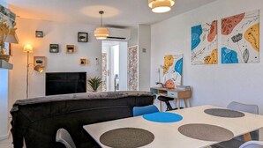 Una sala moderna con un sofá, una mesa de comedor con sillas, y un televisor. Hay obras de arte en las paredes y un escritorio pequeño con una silla.