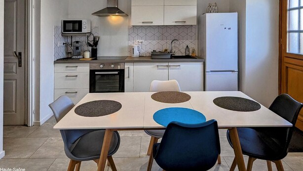 Una cocina moderna con armarios blancos, horno empotrado, microondas y refrigerador. Hay una mesa de comedor con cuatro sillas y una cafetera en la encimera.