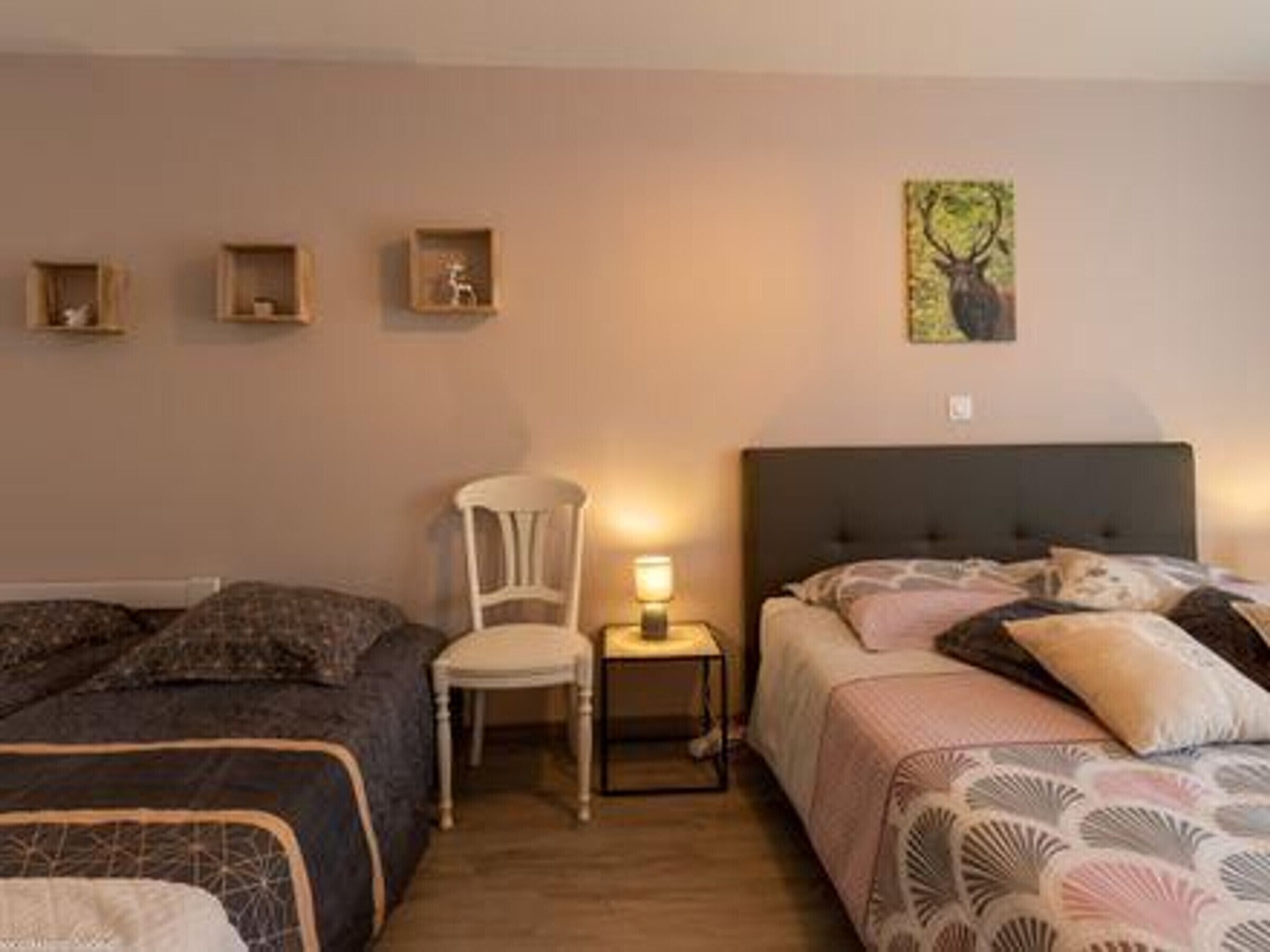 A bedroom with two beds, a chair, a bedside table with a lamp, and wall-mounted shelves with decorative items.