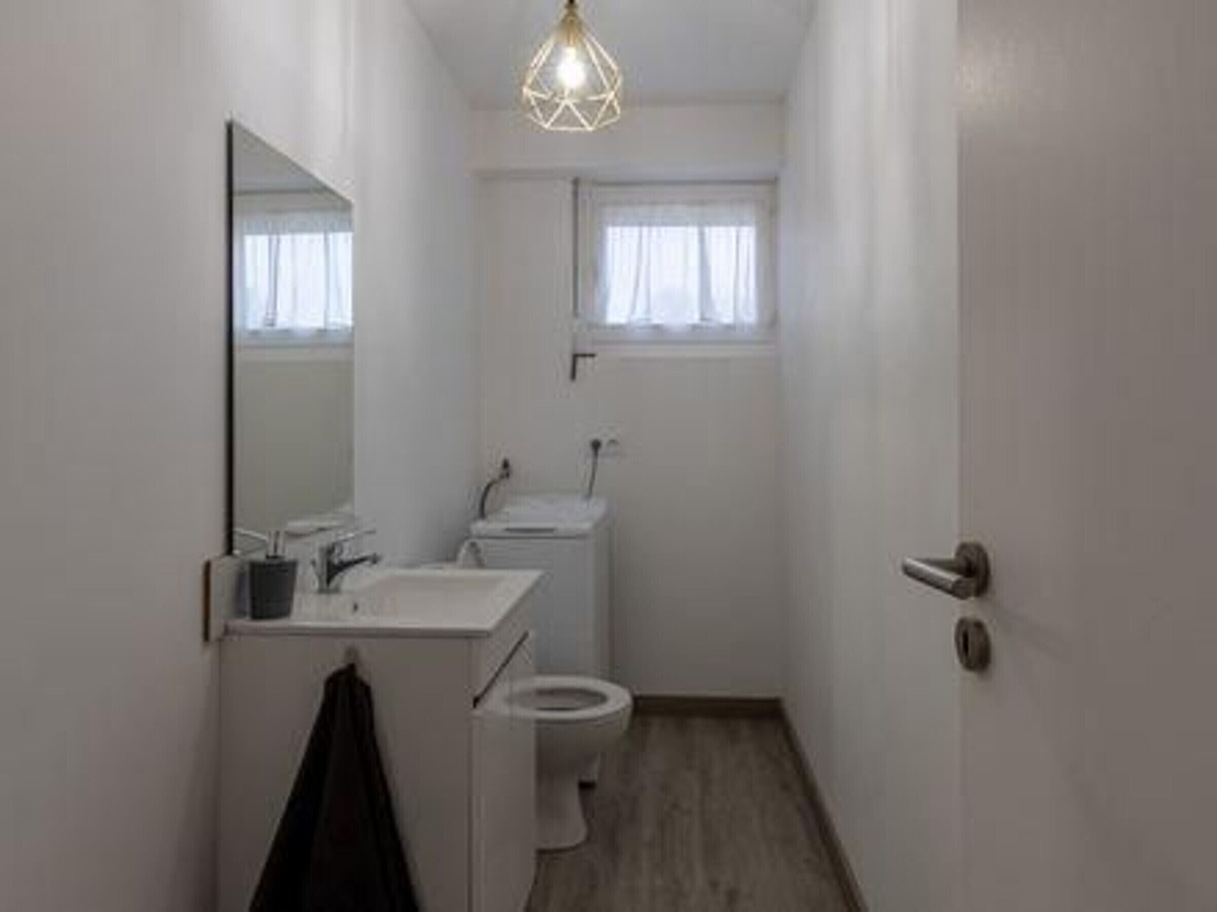 Une petite salle de bain blanche, avec des toilettes, un lavabo et un miroir. Il y a une fenêtre avec des rideaux et un luminaire au plafond.