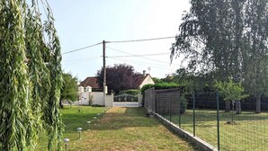 Un chemin mène à une maison à l’extérieur blanc, avec un toit recouvert de tuiles rouges et une cour clôturée.