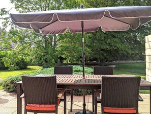 Un patio al aire libre con una mesa y cuatro sillas de mimbre, una sombrilla y un jardín al fondo.