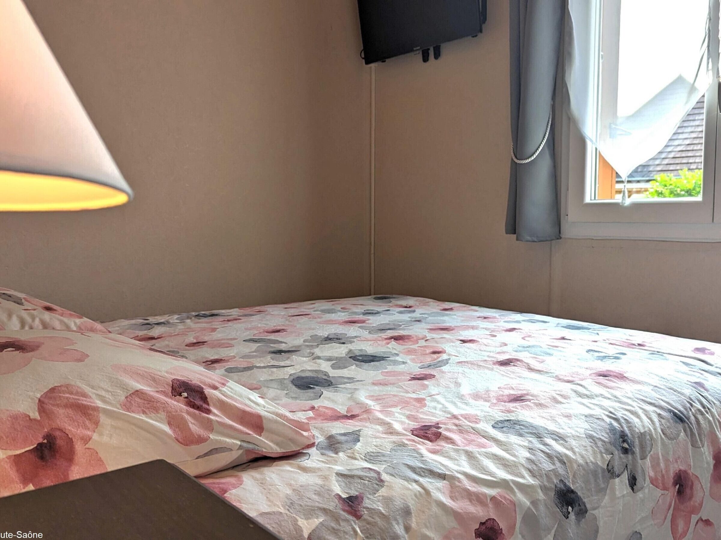 Un dormitorio con cama, lámpara, televisor en la pared y ventana con cortinas.