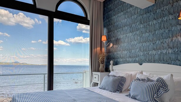 Une chambre avec une grande fenêtre donnant sur un plan d’eau, un lit avec une literie à rayures et un mur texturé derrière le lit.