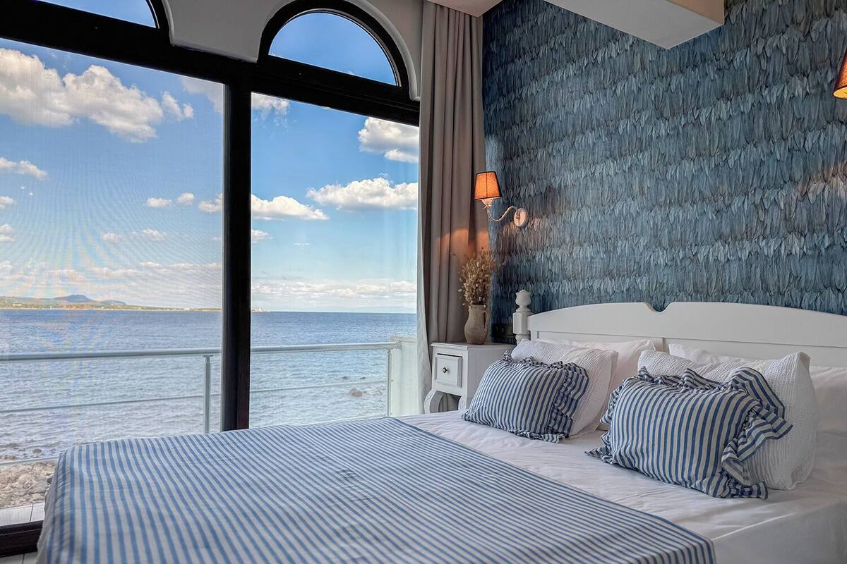 A bedroom with a large window overlooking a body of water, a bed with striped bedding, and a textured wall behind the bed.