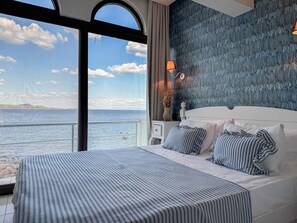 A bedroom with a large window overlooking a body of water, a bed with striped bedding, and a textured wall behind the bed. - Assos Bertiz Hotel (Ayvacik)