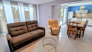 A living room with a sofa, armchair, glass coffee table, and dining area with wooden chairs and a table.