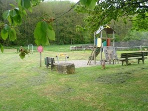 A playground with a slide, climbing structure, and benches in a grassy area with trees in the background.