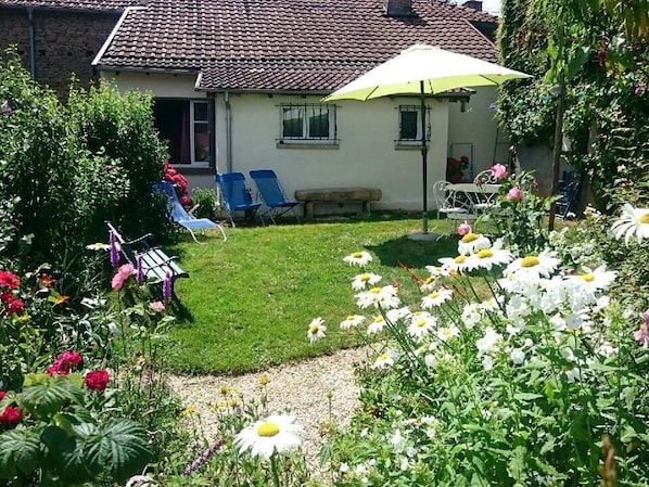 Une maison avec un toit en tuiles, une pelouse et un jardin où poussent différentes sortes de fleurs et de plantes.
