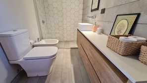 Een moderne badkamer met toilet, bidet en wastafel. Er is een houten wastafelmeubel met een mand en een lijst met een foto aan de muur.