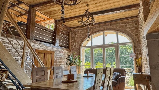 A rustic interior with a stone wall, wooden ceiling, a dining table with chairs, a staircase, and a large arched window. - Woodland Cottage: Comfort in Nature, near the Thousand Lakes Plateau (La Lanterne-et-les-Armonts)