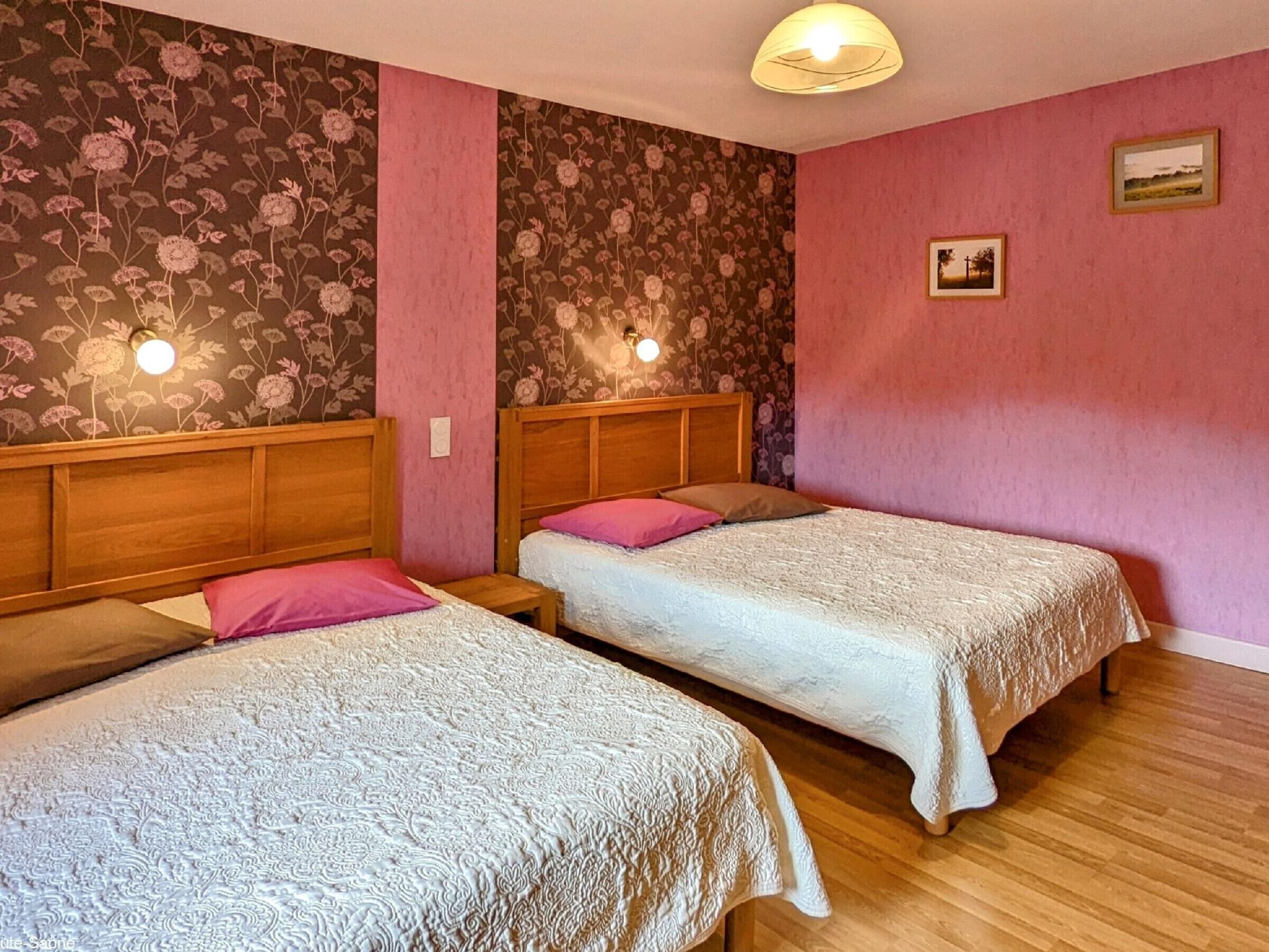 A room with two single beds, wooden headboards, floral wallpaper, pink walls, and a ceiling light fixture.