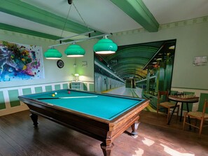 Une table de billard dans une piĂšce aux murs verts, avec un tableau et un escalier menant Ă une mezzanine.