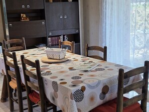 Ein Essbereich mit einem Tisch für vier Personen, umgeben von Holzstühlen. Ein gemustertes Tischtuch bedeckt den Tisch, und ein Korb steht darauf.