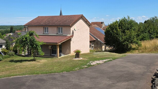 Une maison à deux étages, avec un toit en pente, une cheminée bien visible et un petit porche agrémenté d’une chaise.