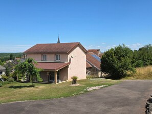 Ein zweistöckiges Haus mit geneigtem Dach, einem sichtbaren Schornstein und einer kleinen Veranda mit einem Stuhl.