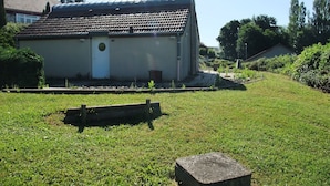 A small, single-story building with a tiled roof, a door, and a small garden area with a bench and a concrete block. - Comfortable Pavilion in the Ognon Valley (Larians-et-Munans)