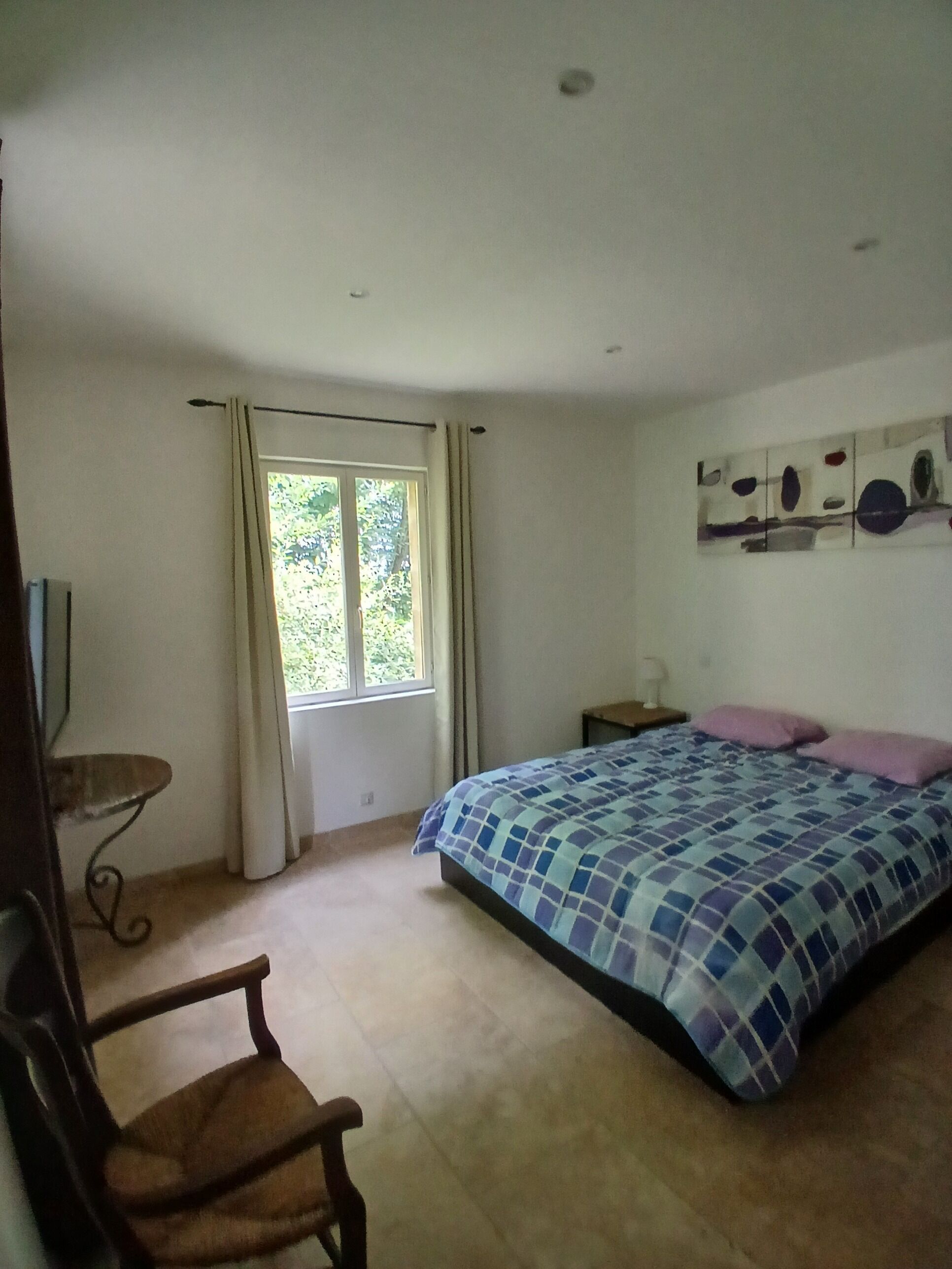 A bedroom with a bed, a chair, a nightstand, a window with curtains, and a wall with a painting.
