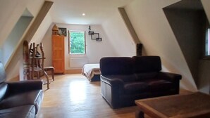 An attic room with a sloped ceiling, wooden flooring, a dark sofa, a wooden coffee table, and a bed with a window view.