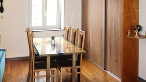Een zithoek met een glazen tafel, houten stoelen, een raam met zonwering, een radiator en een houten kast met decoratieve objecten.