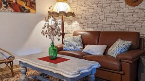 A living room with a brown leather sofa, patterned cushions, a blue table with a red placemat, a green vase with dried flowers, and a stone wall.