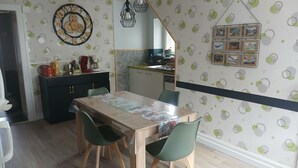 A dining area with a wooden table, green chairs, a kitchen in the background, and a wall clock.