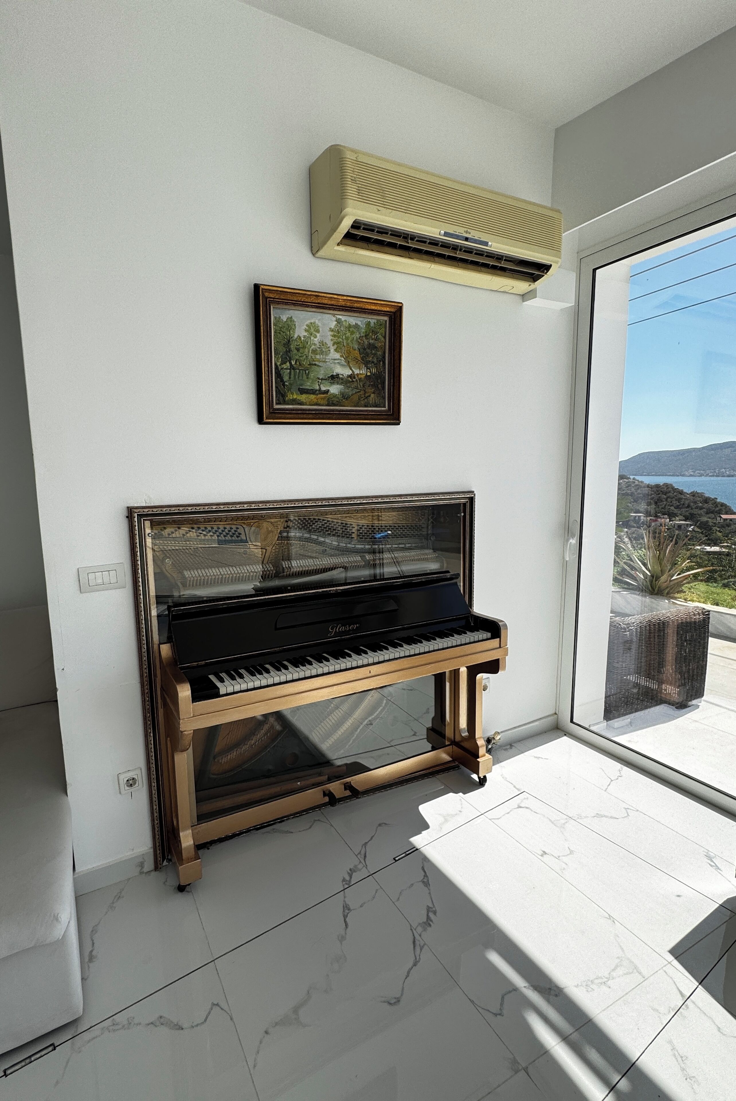 Un pianoforte in una stanza con un'ampia finestra, un quadro appeso al muro e un condizionatore sopra il pianoforte.