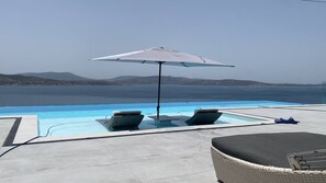 Zona de piscina con sillones de descanso y una sombrilla, con vista a un cuerpo de agua y colinas distantes.