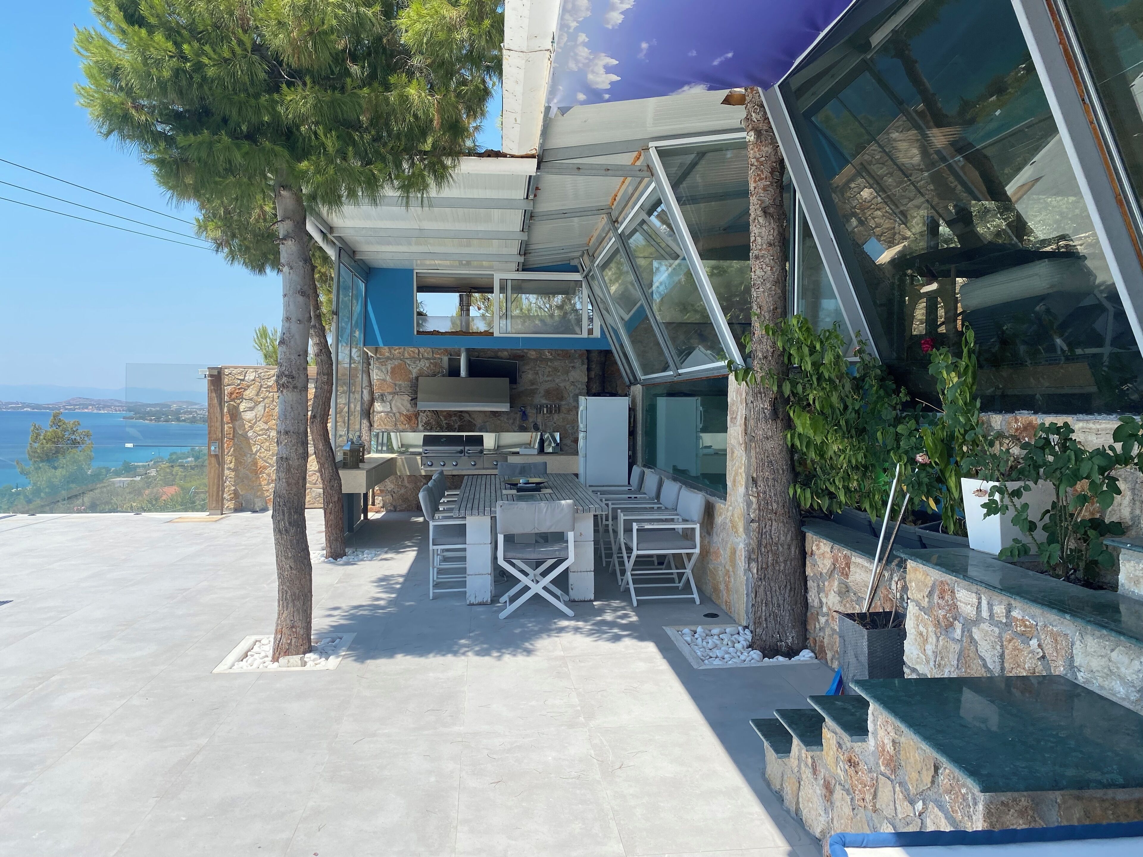 Eine moderne Außenküche mit Steinwand, einem Essbereich mit weißer Möbel und Blick auf das Meer.